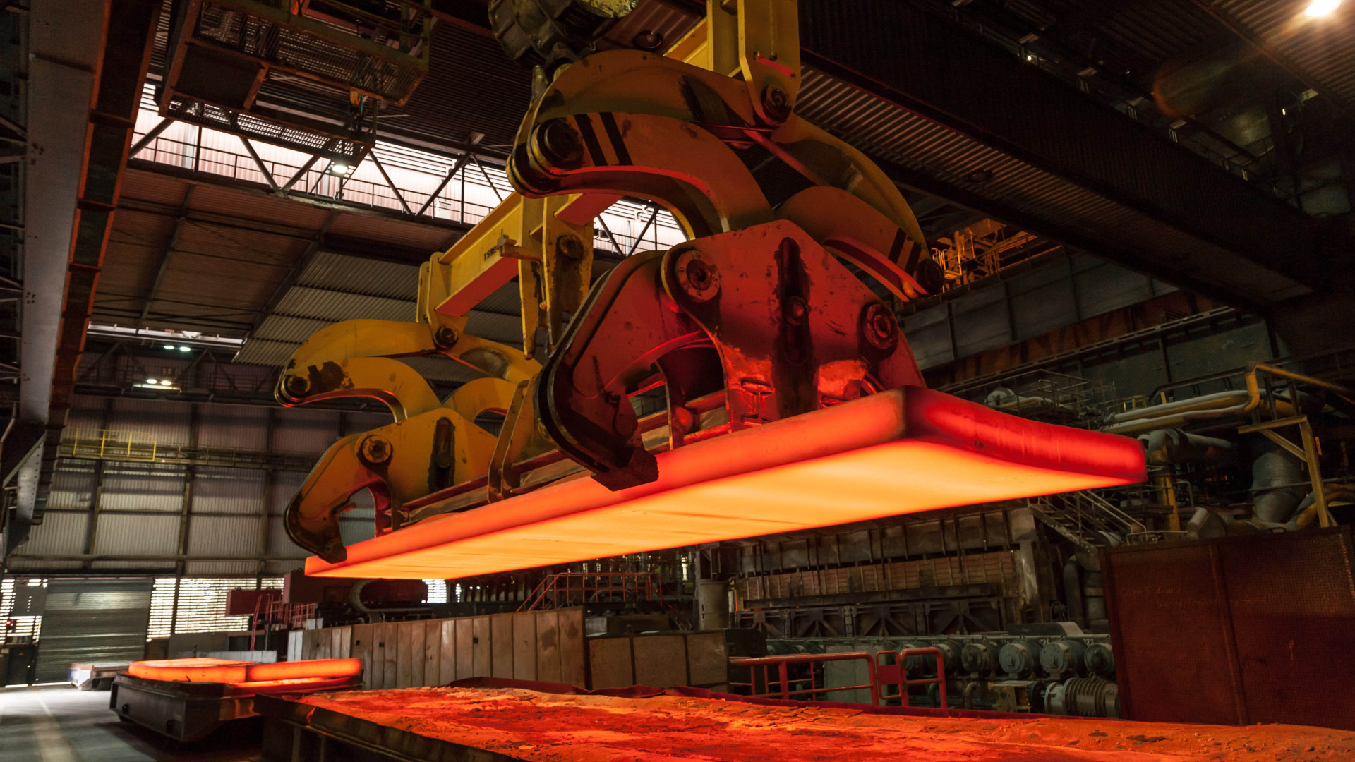 A heavy-duty industrial robotic arm lifts a glowing red-hot steel slab in a steel mill.