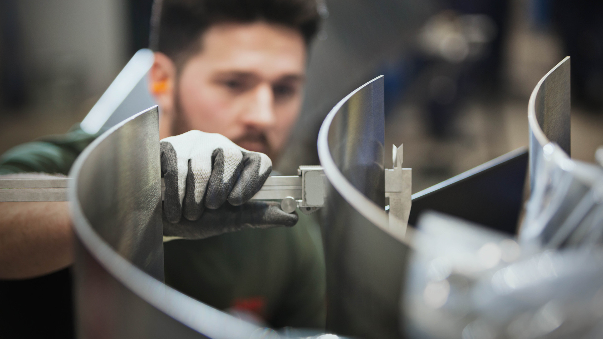 A worker wearing protective gloves uses a caliper to precisely measure a curved metal component in an industrial setting.