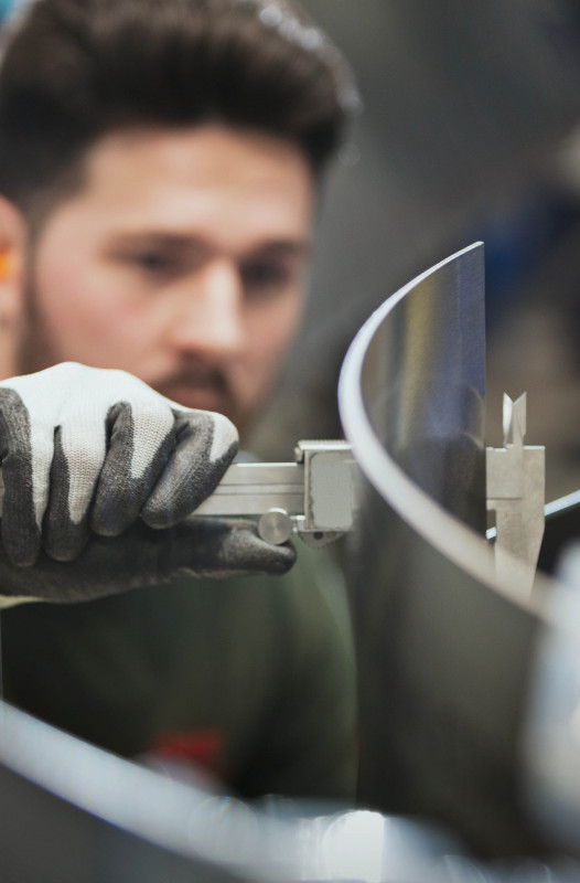 A worker wearing protective gloves uses a caliper to precisely measure a curved metal component in an industrial setting.