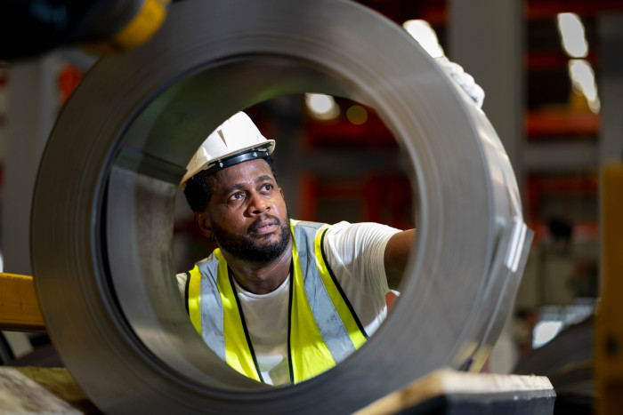 A worker in a high-visibility vest and hard hat inspects a steel coil through its circular opening in an industrial setting.