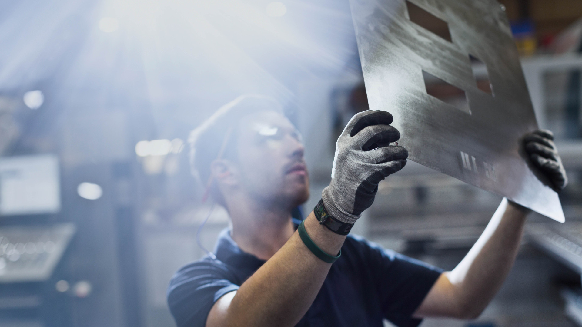 A worker wearing gloves inspects a laser-cut metal sheet under bright industrial lighting.
