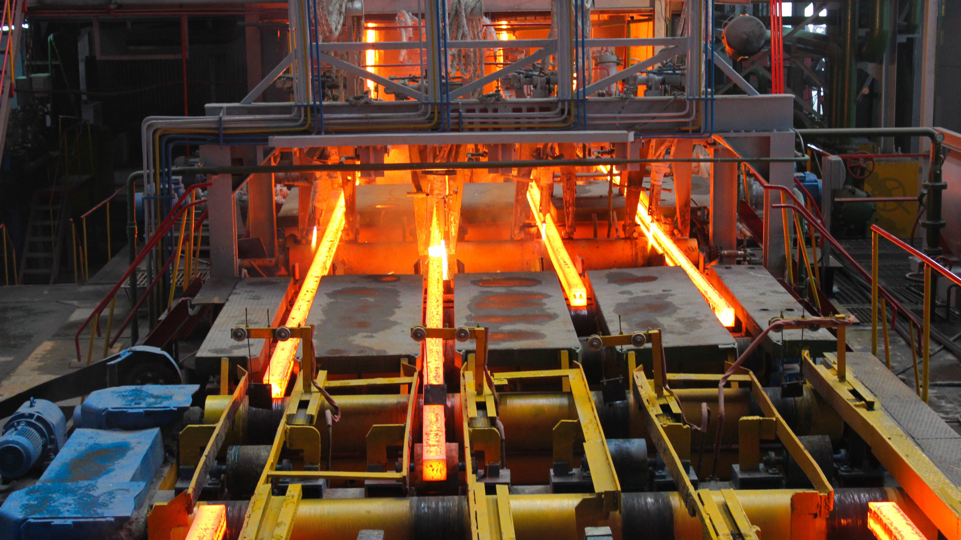 Glowing red-hot steel beams move along a production line in a rolling mill, surrounded by industrial machinery and conveyor systems.